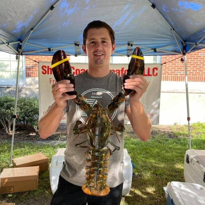 Young man holding a large lobster with rubber bands on its claws, standing under a tent at a seafood market.